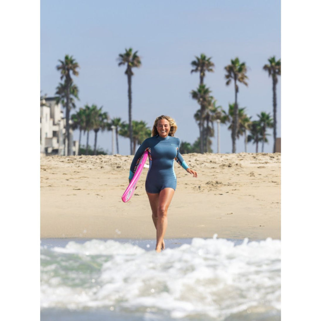 Beach photo of model running into the sea with a surfboard wearing Roxy Swell Series Womens Shorty Wetsuit with long sleeves. Front view. 