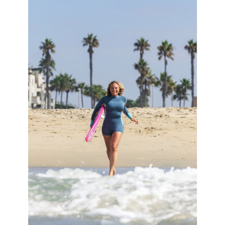 Beach photo of model running into the sea with a surfboard wearing Roxy Swell Series Womens Shorty Wetsuit with long sleeves. Front view. 