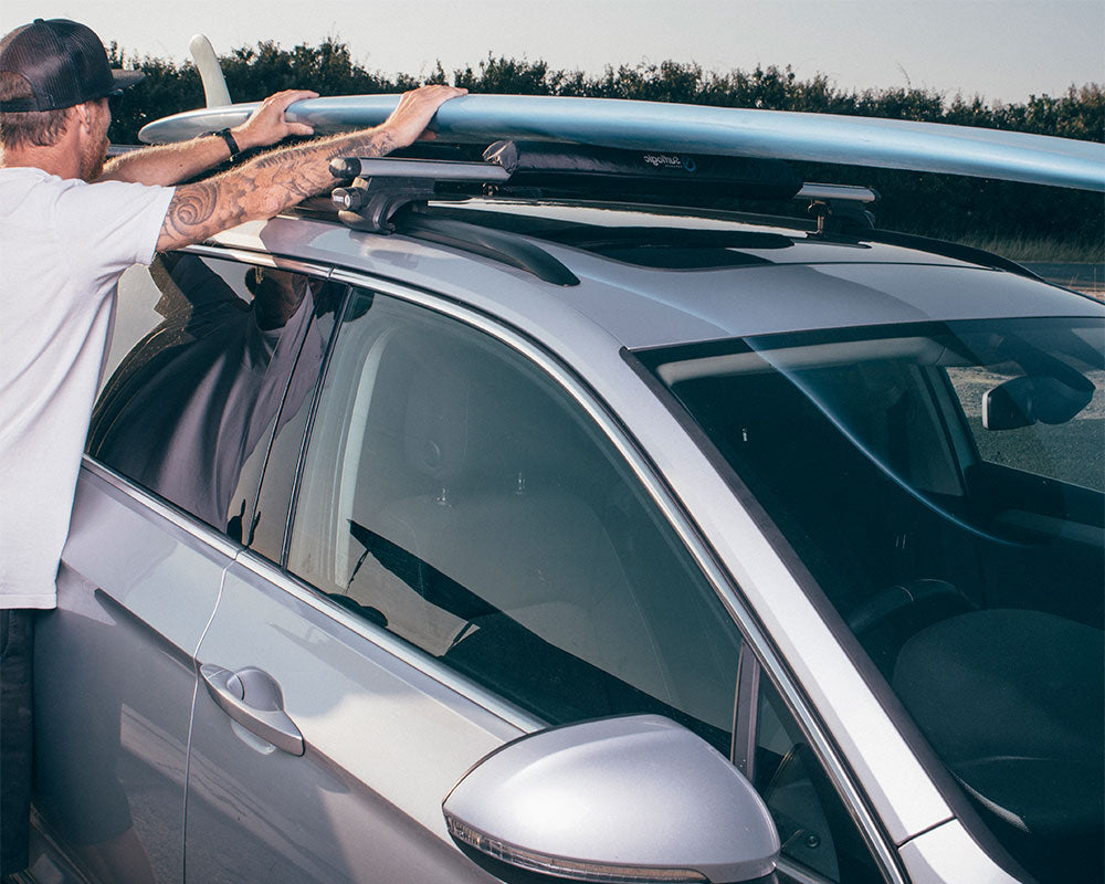 Man loading up a surfboard onto a surflogic roof rack on top of a car. 